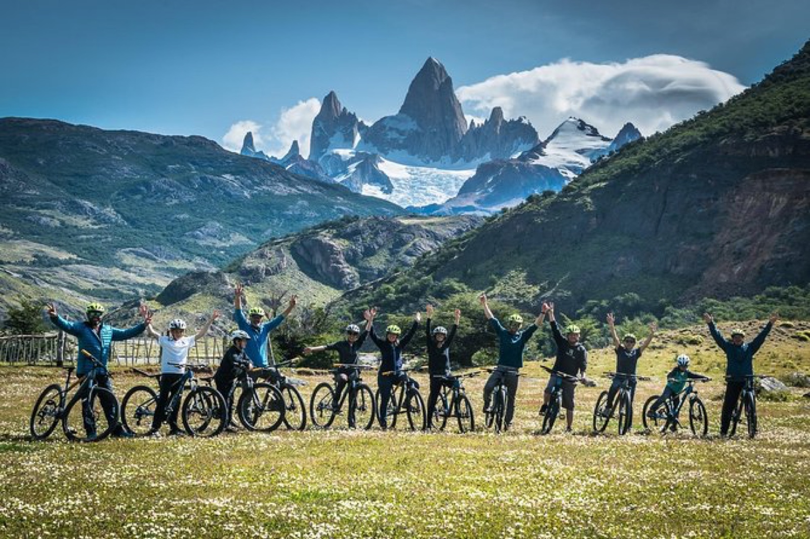 Grupo de viajantes com bicicletas comemorando em campo florido com o Monte Fitz Roy ao fundo